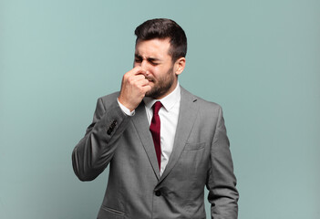 young adult handsome businessman feeling disgusted, holding nose to avoid smelling a foul and...