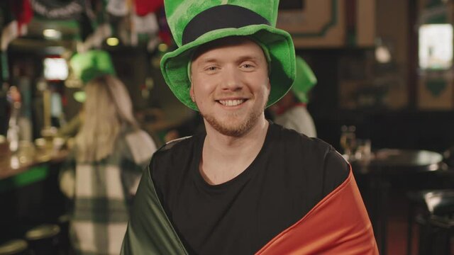 Slow-motion waist-up portrait of smiling caucasian man in green hat and Irish flag on his shoulders looking at camera standing in pub, while his friends dancing in background on St Patricks Day