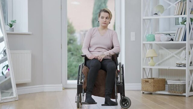 Wide shot portrait of young Caucasian handicapped woman sitting on wheelchair thinking. Sad disabled invalid at home. Concept of disability and hopelessness.