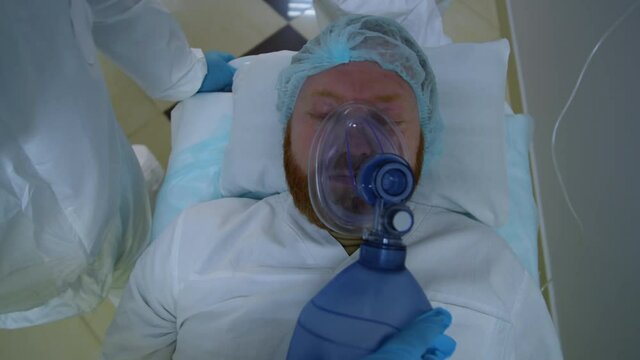 Tilt up shot of paramedics in protective uniform giving bag-valve-mask ventilation and IV fluid to covid-19 patient, pushing gurney and running along hospital hallway