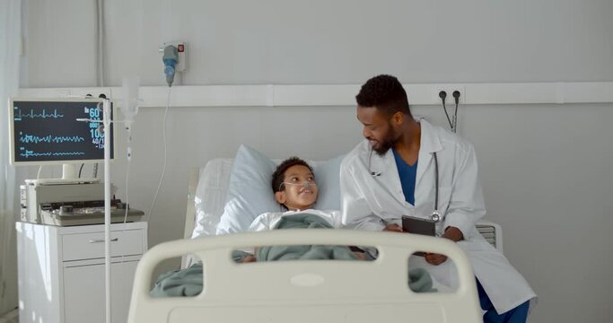 Young african doctor sitting on hospital bed and talking to preteen boy patient