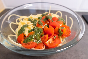 Glass bowl full of spaghetti and cut cherry tomatoes with green basil pesto on top