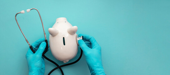 Health care cost. Doctor in surgical gloves holding a white piggy bank and stethoscope - obrazy, fototapety, plakaty