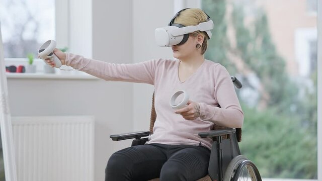Active young paraplegic woman using virtual reality headset sitting on wheelchair indoors. Portrait of positive Caucasian handicapped using VR headset and motion controllers gaming online.