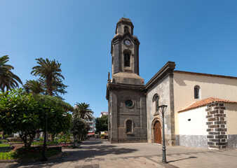 A church in the center of the Canary town of Puerto de La Cruz on the island of Tenerife. It's a sunny autumn day with blue skies. In front of the church is a small park with trees.