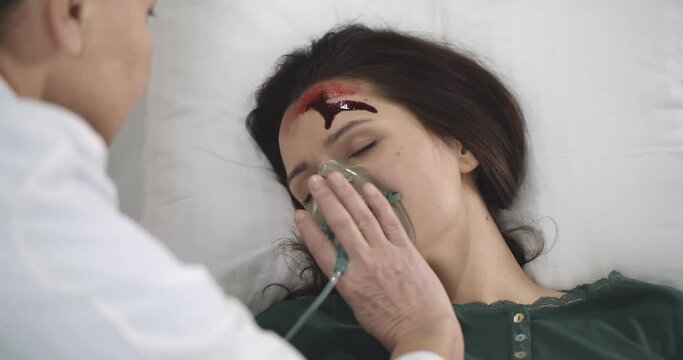 Doctor pressing oxygen mask to woman face lying unconscious on bed with bleeding wound on forehead