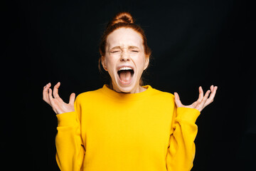 Close-up portrait of excited angry young woman in yellow sweater screaming with closed eyes on isolated black background. Pretty redhead lady model emotionally showing facial expressions, copy space.