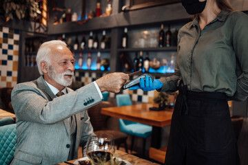 Waitress serves senior gentleman. She holds a card reader while businessman uses his credit card to pay bill. She wears a protective mask as part of security measures against the Coronavirus pandemic.