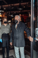 Naklejka na ściany i meble Senior businessman walks out of a restaurant. Young waitress kindly sees him off. They are wearing protective face mask as protection against virus pandemic.