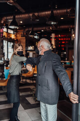 Fototapeta na wymiar Senior businessman walks out of a restaurant. Young waitress kindly sees him off. They are wearing protective face mask as protection against virus pandemic.