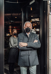 Fototapeta na wymiar Senior businessman walks out of a restaurant. Young waitress kindly sees him off. They are wearing protective face mask as protection against virus pandemic.