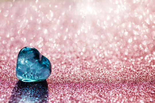 Blue glass heart on the shining pink bokeh background. Focus on the left bottom on the heart. Valentines day. Light shines through the heart. Greeting card, copy space.