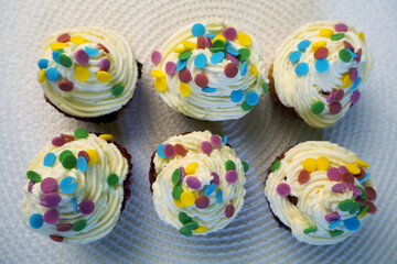 six yogurt muffins with red currants, spread with white cream of cream and mascarpone and decorated with colorful round splashes, lie on a white table stand with a view from above . homemade cakes for