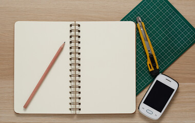 Blank Spiral Note Pad, cutter, cellphone and Pencil on Wood Background.