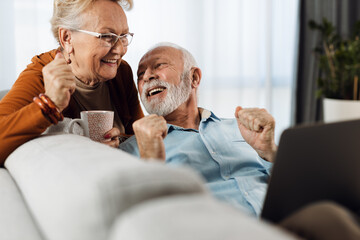 Fototapeta premium Cheerful senior couple relaxing on sofa in the living room and using laptop