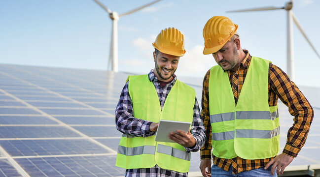 Men Working At Solar Power Station With Digital Tablet - Solar Panels With Wind Turbines In Background - Green Energy Renewable Concept