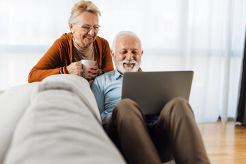 Cheerful senior couple relaxing on sofa in the living room and using laptop