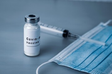 Vaccine ampoule with COVID-19 inscription, injection syringe and medical protective mask on a gray...