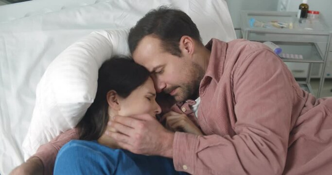 Husband hugging and comforting crying sick woman lying together in hospital bed