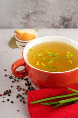 hot homemade chicken broth with collagen and amino acids in red bowl with red napkin and green onions, black peas, chopped white onions and carrots on gray background