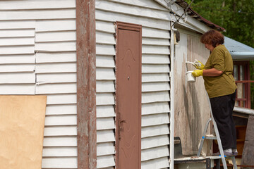 Fototapeta premium A middle-aged woman in T-shirt outdoors paints a wooden building with a brush with white paint