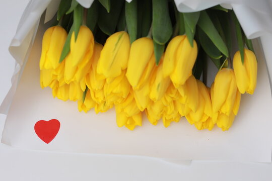 Beautiful Big Bouquet Of Yellow Tulips In A White Package And Red Heart Confetti On White Table Close Up. Congratulation. Festive Background. Copy Space.