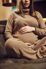 Cute pregnant woman sitting on sofa at her home and cuddling tummy.