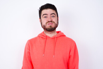 young Caucasian bearded man wearing pink hoodie against white wall puffing cheeks with funny face. Mouth inflated with air, crazy expression.