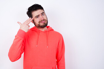 Unhappy young Caucasian bearded man wearing pink hoodie against white wall imitates gun shoot makes suicide gesture keeps two fingers on temples.