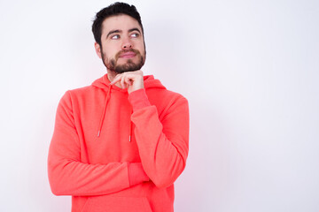 Dreamy young Caucasian bearded man wearing pink hoodie against white wall with pleasant expression, looks sideways, keeps hand under chin, thinks about something pleasant.