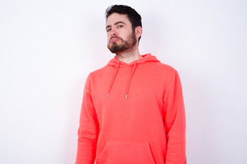 young Caucasian bearded man wearing pink hoodie against white wall with snobbish expression curving lips and raising eyebrows, looking with doubtful and skeptical expression, suspect and doubt.