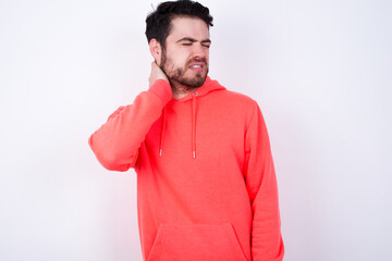 young Caucasian bearded man wearing pink hoodie against white wall suffering from back and neck ache injury, touching neck with hand, muscular pain.
