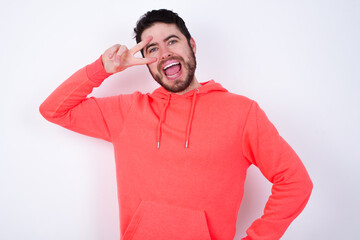 young Caucasian bearded man wearing pink hoodie against white wall making v-sign near eyes. Leisure, coquettish, celebration, and flirt concept.