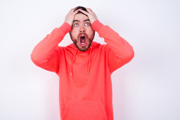 Horrible, stress, shock. Portrait emotional crazy young Caucasian bearded man wearing pink hoodie against white background clasping head in hands. Emotions, facial expression concept.