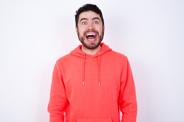 Fototapeta premium Surprised young Caucasian bearded man wearing pink hoodie against white background, shrugs shoulders, looking sideways, being happy and excited. Sudden reactions concept.