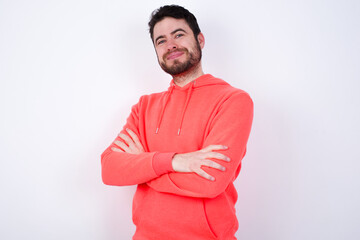 Waist up shot of  self confident young Caucasian bearded man wearing pink hoodie against white wall has broad smile, crosses arms, happy to meet with colleagues.
