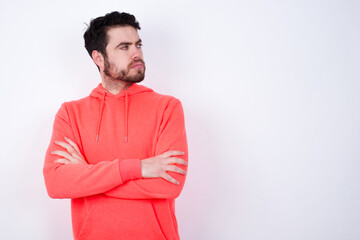 Image of upset young Caucasian bearded man wearing pink hoodie against white wall with arms crossed. Looking with disappointed expression aside after listening to bad news.