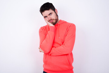 Very bored young Caucasian bearded man wearing pink hoodie against white wall holding hand on cheek while support it with another crossed hand, looking tired and sick.