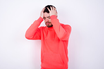 young Caucasian bearded man wearing pink hoodie against white wall holding head with hands, suffering from severe headache, pressing fingers to temples