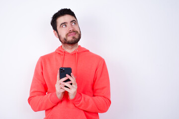 Portrait of young Caucasian bearded man wearing pink hoodie against white wall with dreamy look, thinking while holding smartphone. Tries to write up a message.
