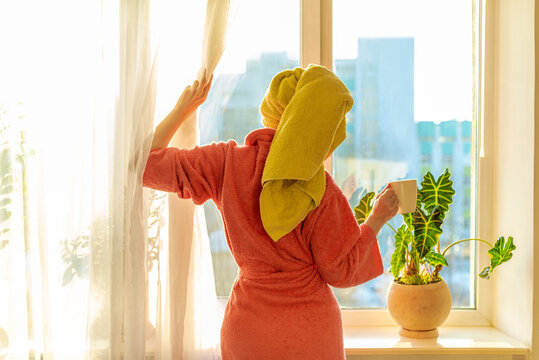 Back View Of A Woman In A Bathrobe And A Turban Towel On Her Head Pulling Back The Curtain