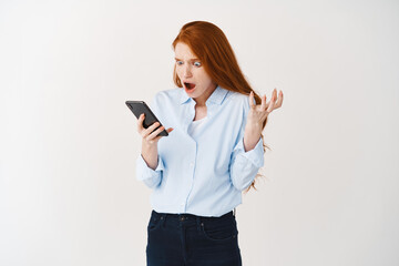 Shocked and confused young businesswoman with red hair staring at smartphone screen startled, cant understand what happened, read bad news, white background