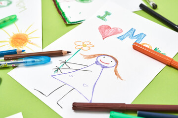 The best gift from child. Mothers day greeting card with text I love mom, child drawing and colored felt tip pens lying on green table