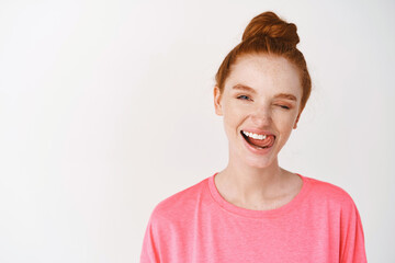 Girl with ginger hair combed in messy bun, winking joyfully and showing perfectly white smile,...