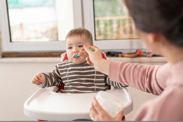 Young mother feeding her cute adorable baby boy, whom sitting on the highchair, with a spoon. Family, food, child, eating, parenthood and motherhood concept. Lifestyle and real people concept.