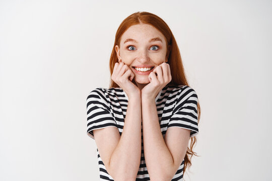 Image of extremely happy and surprised young woman with red hair, touching face and smiling amazed, receiving surprise gift, white background