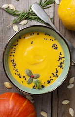 Pumpkin cream soup with black cumin, rosemary and seeds in a green bowl. Top view of autumn soup, ripe pumpkins, rosemary twigs, spoon on a wooden table in rustic style