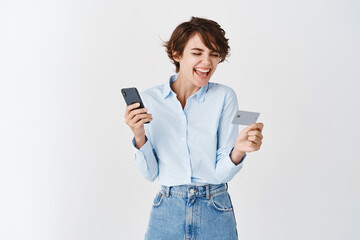 Naklejka premium Cheerful laughing woman looking at plastic credit card and using smartphone, order online, shopping in internet, standing against white background
