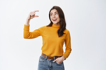 Smiling young woman showing small size hand and looking at little thing with happy and satisfied face expression, standing in casual clothes against white background