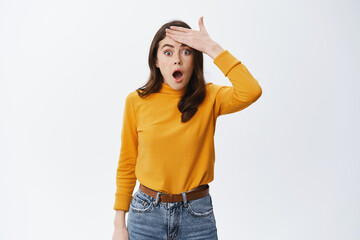 Forget remember. Shocked woman slap forehead and gasping startled at camera, standing in casual yellow sweater and jeans against white background